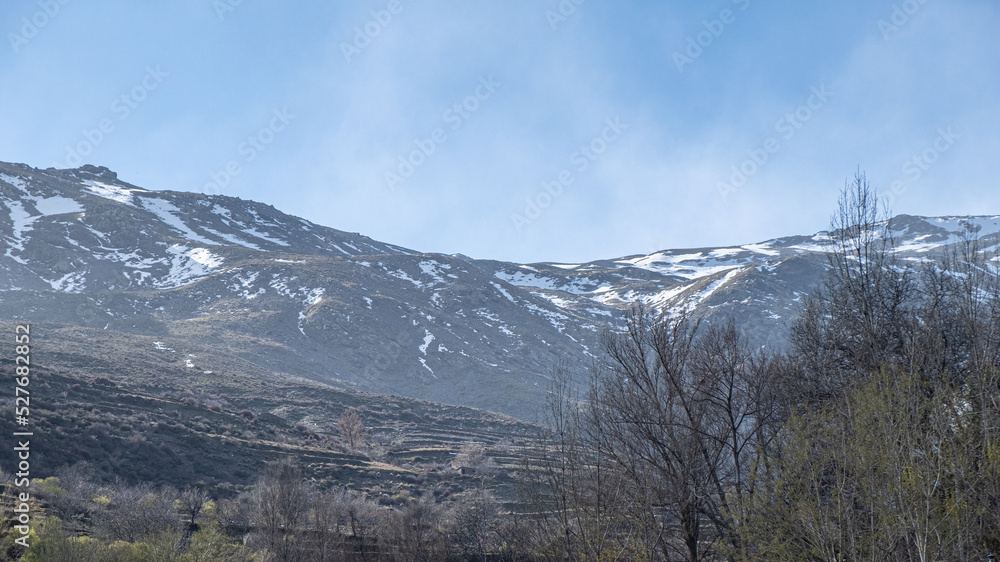 Bottom-up view to rocky mountains partly covered by snow with clear blue sky in background and empty winding paved road along clay cliff partially overgrown in the foreground. Bottom-up view to rocky mountains partly covered by snow with clear blue sky in background and empty winding paved road along clay cliff partially overgrown in the foreground.