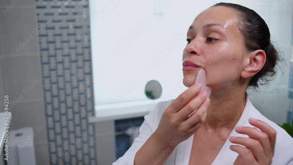 Middle aged pretty woman in bathrobe applies a moisturizing serum to her face and using Gua Sha rose quartz scraper, performs smoothing rejuvenation massage. Alternative rejuvenating physical therapy Middle aged pretty woman in bathrobe applies a moisturizing serum to her face and using Gua Sha rose quartz scraper, performs smoothing rejuvenation massage. Alternative rejuvenating physical therapy