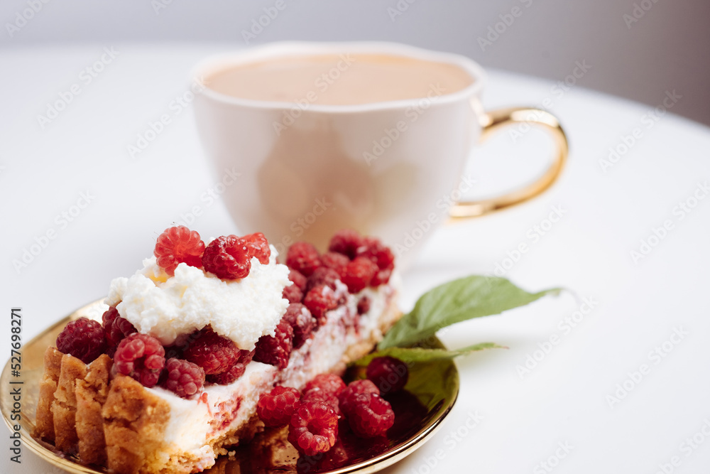Delicious piece of cake with fresh raspberries and blurred cup of aromatic coffee in background. Blurred white background. Sweet concept. Excellent image for dessert banners and advertisements. Delicious piece of cake with fresh raspberries and blurred cup of aromatic coffee in background. Blurred white background. Sweet concept. Excellent image for dessert banners and advertisements.
