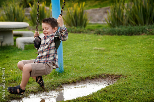 Niño feliz en el parque en el columpio jugando divertido colgado arriba de un charco de agua lleno de lodo al aire libre disfrutando de un hermoso día