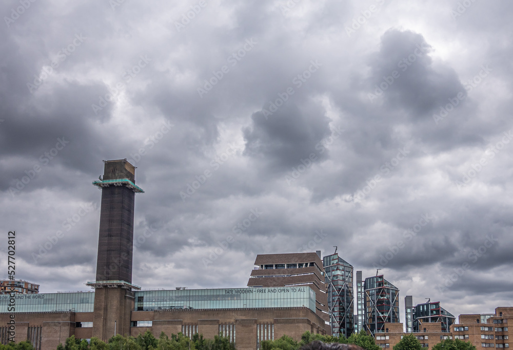 London, England, UK - July 6, 2022: From Thames River. On south shore, Tate modern art museum under heavy cloudscape with its tower and glass upper level