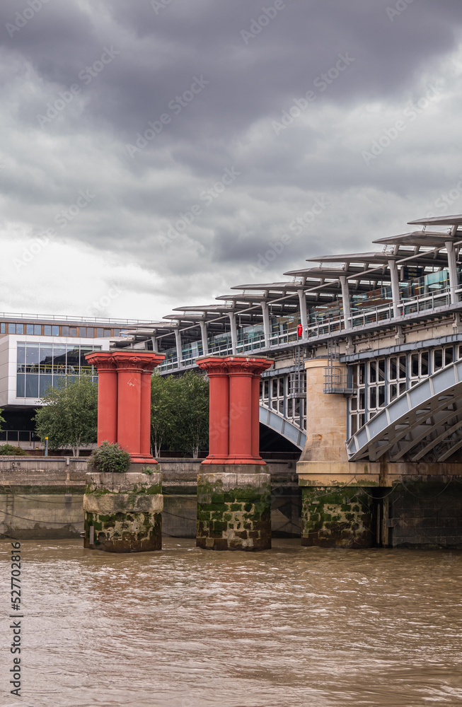 Naklejka premium London, England, UK - July 6, 2022: From Thames River. Oxblood red pillars protecting Blackfriars railway bridge NW shore under thick gray cloudscape.