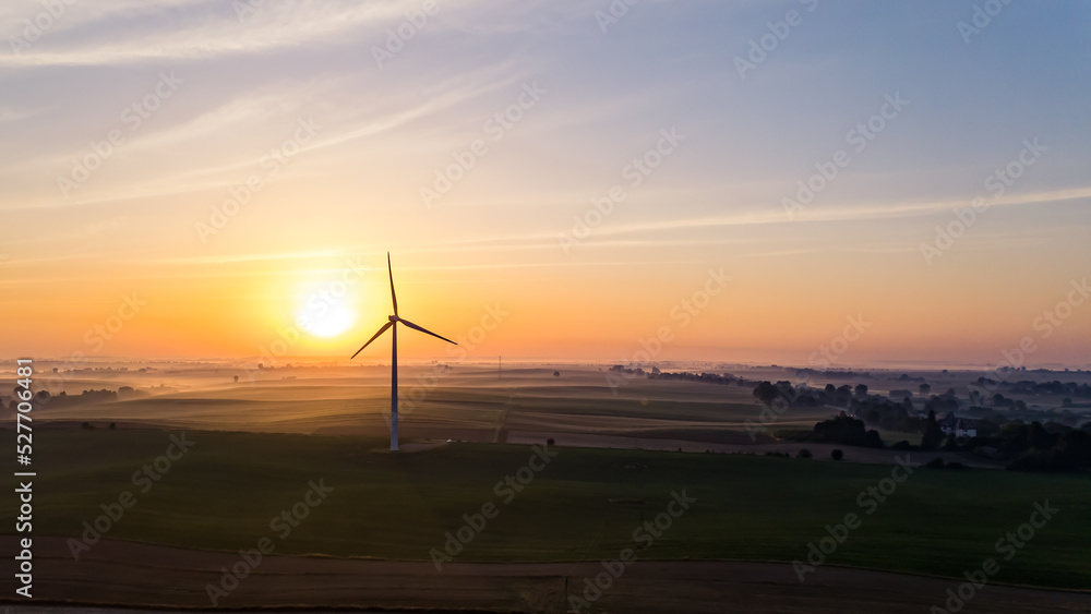 Bright orange rising sun over farmfields with silhouette of wind turbine and trees. Colorful sky. Environmentally friendly energy. Horizontal shot. High quality photo