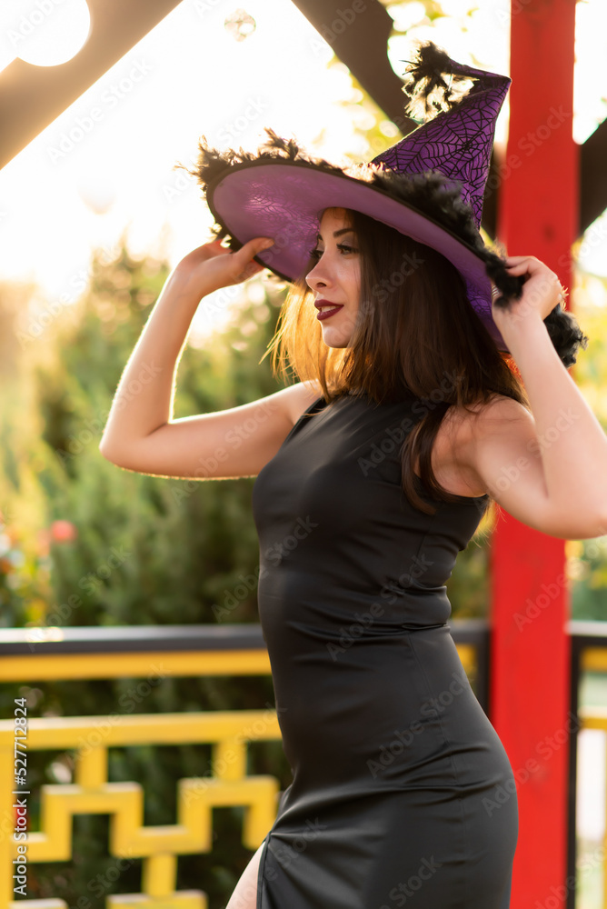 Beautiful girl in a black dress and a witch's hat posing in a gazebo in the sun