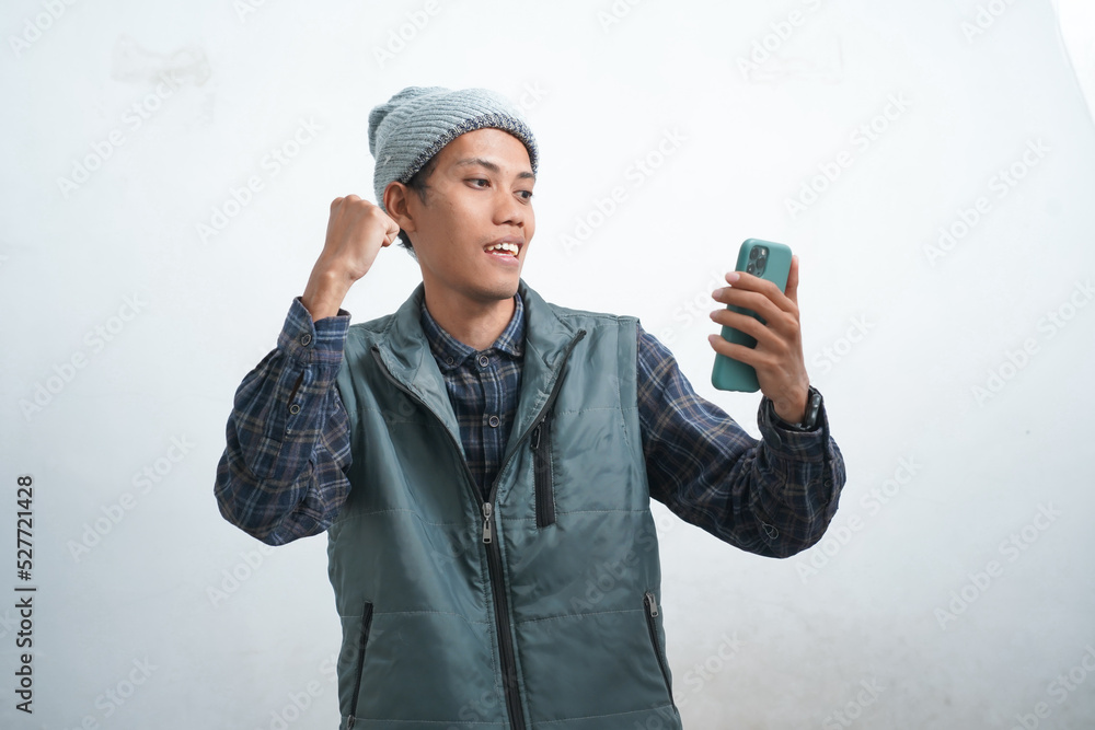 Asian young man wearing dark blue vest and beanie, isolated on white background looks happy looking at his phone screen. Asian young man wearing dark blue vest and beanie, isolated on white background looks happy looking at his phone screen.