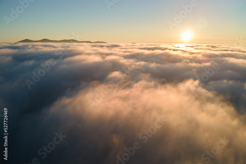 Aerial view of colorful sunrise over white dense fog with distant dark silhouettes of mountain hills on horizon