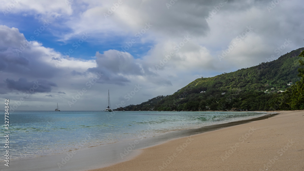 Sandy beach on a tropical island. The turquoise ocean is calm. Yachts are visible in the distance. A green hill against a background of blue sky and clouds. Seychelles. Mahe. Beau Vallon