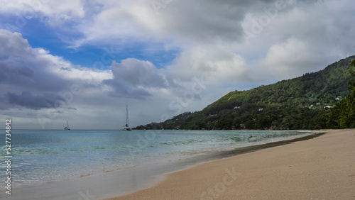 Fototapeta Naklejka Na Ścianę i Meble -  Sandy beach on a tropical island. The turquoise ocean is calm. Yachts are visible in the distance. A green hill against a background of blue sky and clouds. Seychelles. Mahe. Beau Vallon