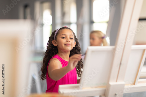 Cute biracial elementary schoolgirl painting on easel during drawing class in school