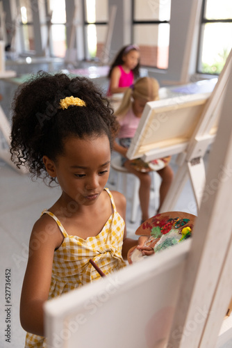 Biracial elementary schoolgirl holding paint palette while painting on easel during drawing class