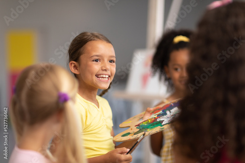 Happy caucasian elementary schoolgirl sitting by multiracial female classmates during drawing class