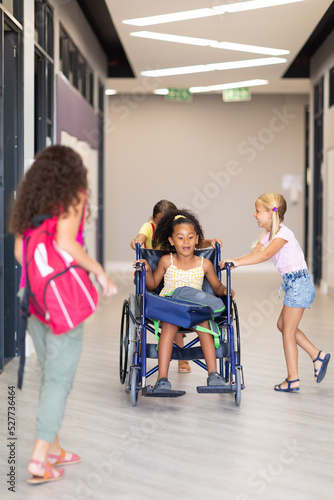 Multiracial elementary schoolgirls holding female classmate wheelchair in corridor