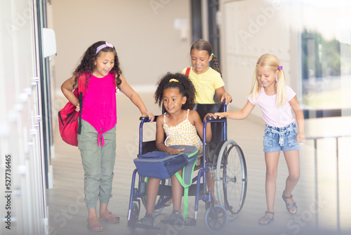 Smiling multiracial elementary schoolgirls assisting female biracial classmate sitting on wheelchair