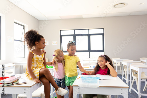 Multiracial elementary schoolgirls looking at female classmate reading book at desk in classroom