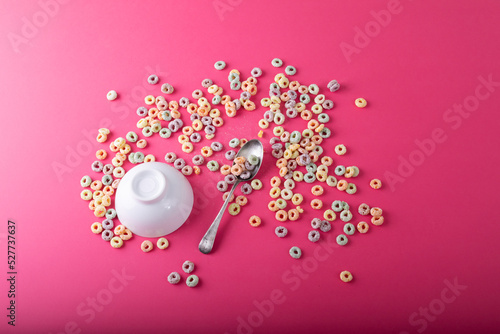 High angle view of upside down bowl and spoon with ring shaped breakfast cereal over pink background