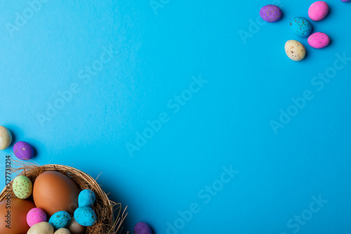 Overhead view of easter eggs with colorful candies in nest on blue background with copy space