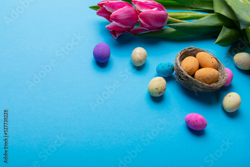 Colorful easter candies in small nest with pink tulips on blue background with copy space