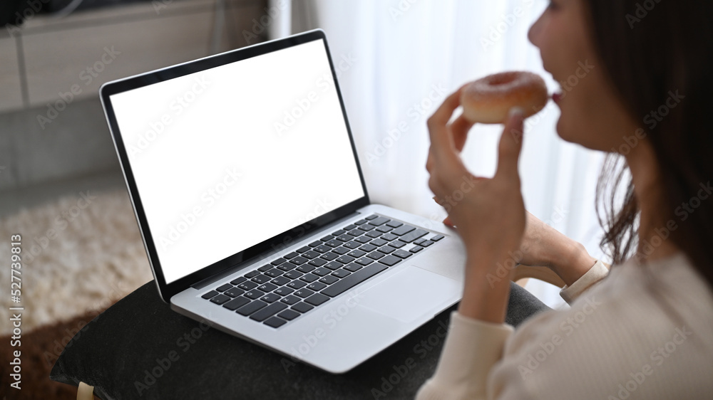 Over shoulder view of young woman eating donut and browsing internet, shopping online on laptop at home Over shoulder view of young woman eating donut and browsing internet, shopping online on laptop at home