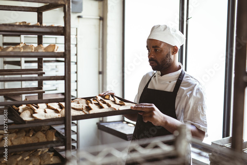 African american mid adult male baker removing baked bread tray from rack in bakery