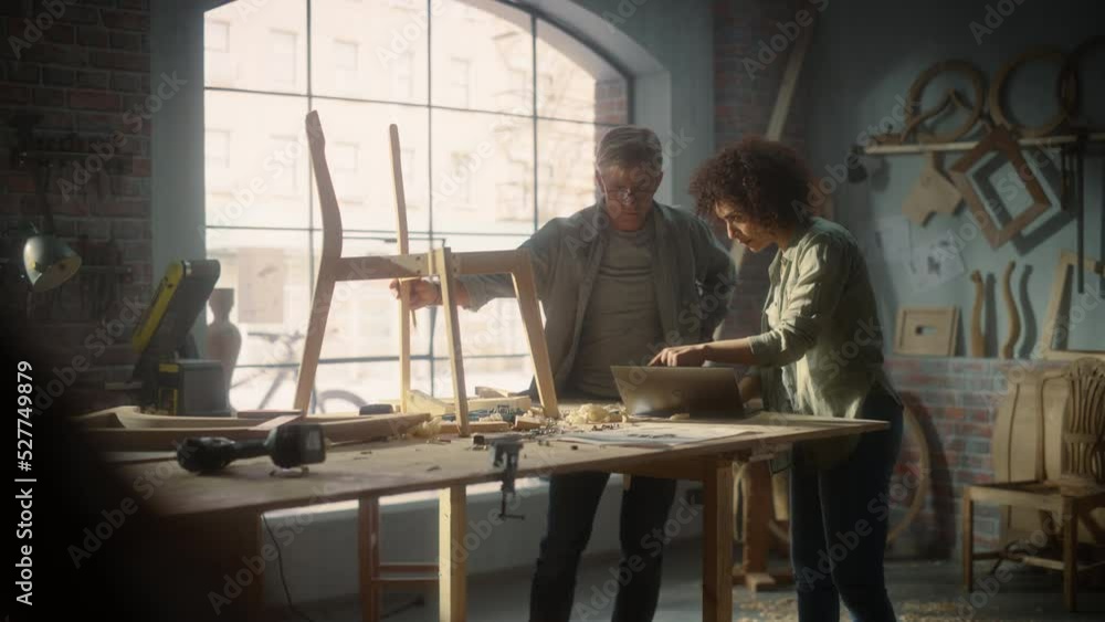 Small Business Owners of a Furniture Workshop Using Laptop Computer and Discussing the Design of a New Wooden Chair. Middle Age Carpenter and a Young Female Apprentice Working in Loft Studio. Small Business Owners of a Furniture Workshop Using Laptop Computer and Discussing the Design of a New Wooden Chair. Middle Age Carpenter and a Young Female Apprentice Working in Loft Studio.