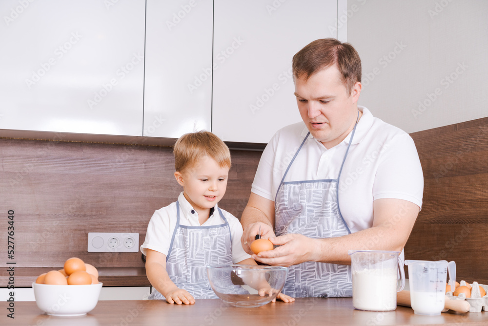 Dad and son in kitchen, father teaches child to break eggs into a bowl. Cooking together at home. The son helps his parents. Happy father's day time Dad and son in kitchen, father teaches child to break eggs into a bowl. Cooking together at home. The son helps his parents. Happy father's day time