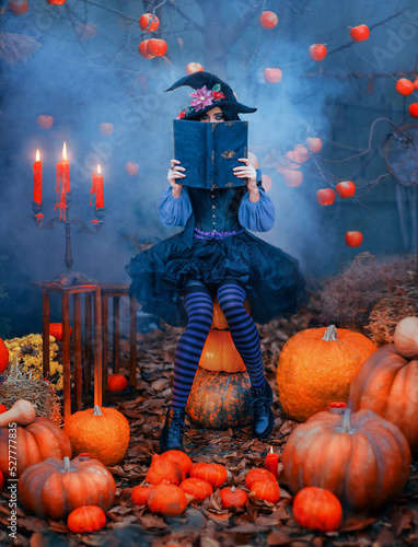 Slika na platnu Fantasy woman halloween witch holds magic book in hands hides face sits on orange pumpkin