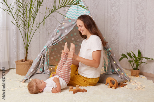 Portrait of happy smiling delighted mother wearing white t shirt sitting in peetee tent and having fun with her daughter lying on floor with raised leg, positive family spending weekend. photo