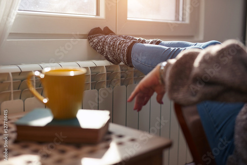 Woman heating feet on a chilly winter day, energy and gas crisis, cold room, heating problems.