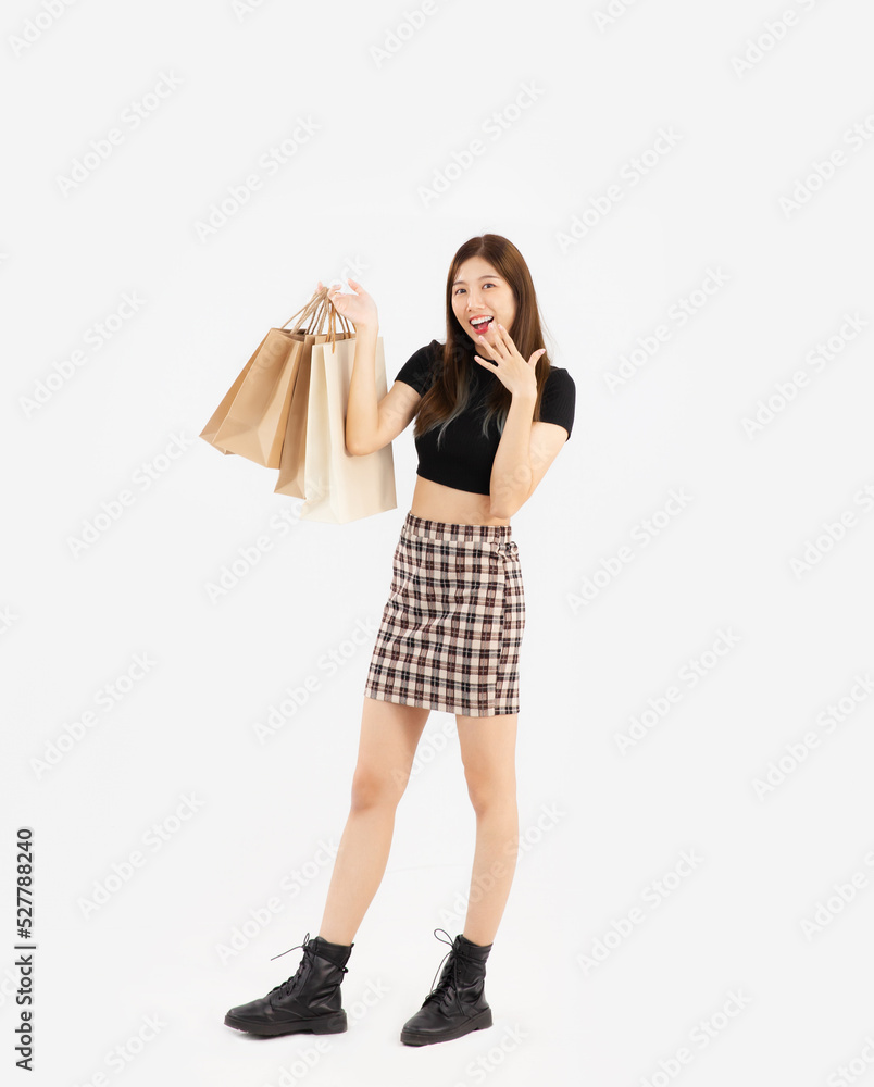 Young asian woman in black crop top t-shirt striped skirt and ankle boots carrying brown paper shopping bags posing surprised on white background, full body.