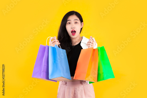 Young asian woman long hair carrying green, blue orange and purple shopping paper bag posing wow and excited on yellow background.