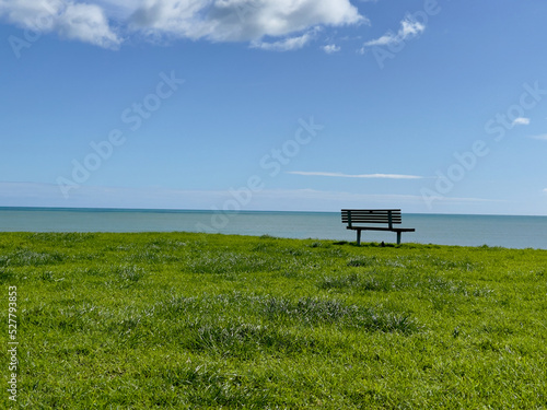 A sunny day looking out over the ocean in New Zealand with an empty park bench in the mid-ground