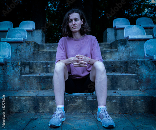 european girl in black shorts pink t-shirt sitting in the stadium cloudy weather cold sad depression upset seriousness