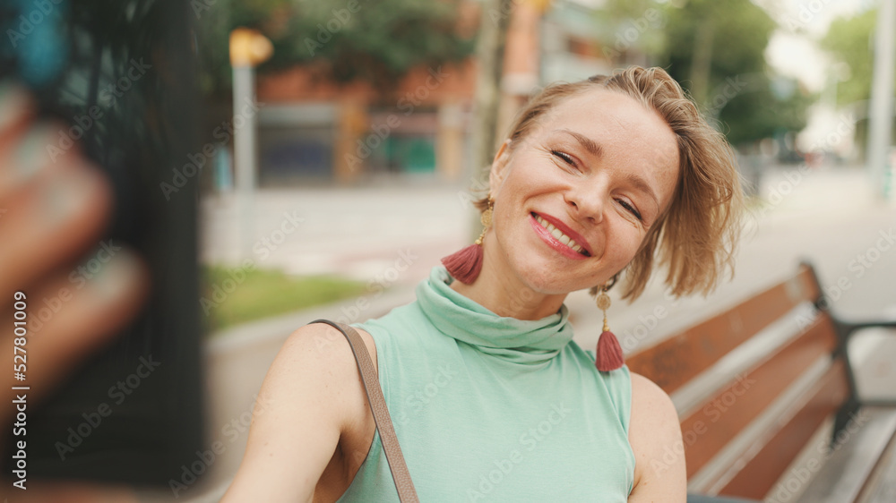 Beautiful happy smiling woman with short blond hair in casual wear sits on bench, makes video call on cellphone Beautiful happy smiling woman with short blond hair in casual wear sits on bench, makes video call on cellphone