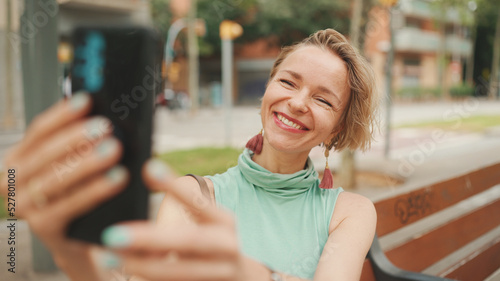 Beautiful happy smiling woman with short blond hair in casual wear sits on bench, makes video call on cellphone