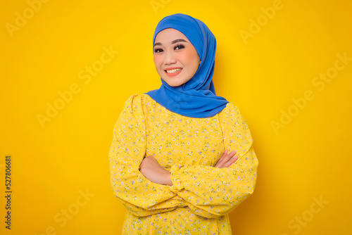 Smiling young Asian woman in casual dress standing with crossed arms isolated on yellow background