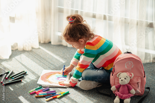 A cute little girl sits on the floor in the rays of the sun and draws a rainbow with colorful markers. She is dressed in a colored jumper. Creation. Childhood.