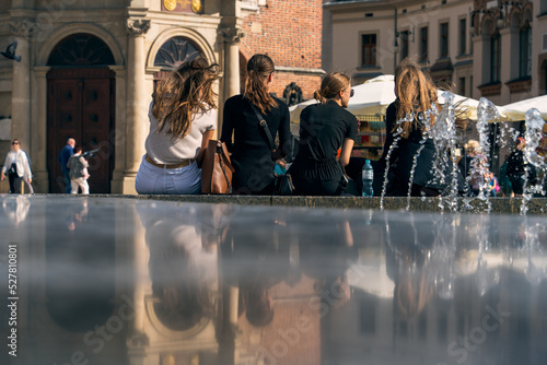 Four friends are sitting near the fountain on a sunny day, young female students are resting by the fountain after class, reflections on the marble surface, the charm of youth