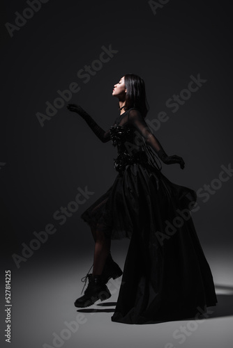 side view of asian young woman in black gothic dress and gloves levitating on dark grey.