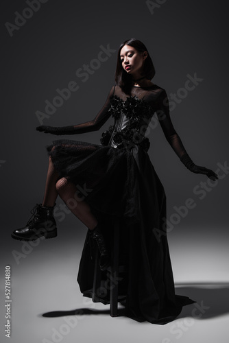 full length of asian young woman in black gothic dress and gloves sitting on high chair on dark grey.