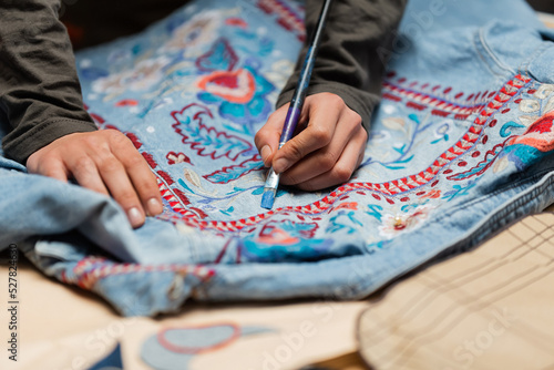 Fototapeta Naklejka Na Ścianę i Meble -  Cropped view of african american designer painting on denim jacket near blurred sewing pattern.