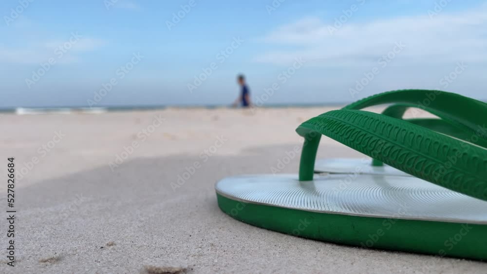 Close up green and white rubber slippers on sandy beach at beachfront with blue sky landscape ocean waves on sunny day. Blurred male walking on beach in background Close up green and white rubber slippers on sandy beach at beachfront with blue sky landscape ocean waves on sunny day. Blurred male walking on beach in background