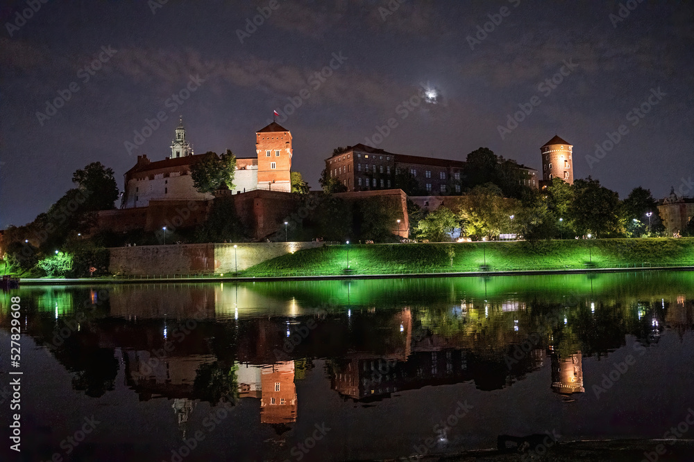 KRAKOW, POLAND: A view of Wawel Royal Castle in cracow at night from across the river Vistula illuminated during full moon with reflection on a calm water.