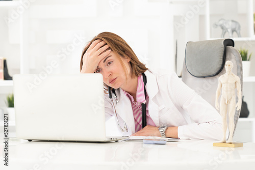 Close up portrait of tired female doctor sitting at the desktop and working on laptop in the office of modern clinic