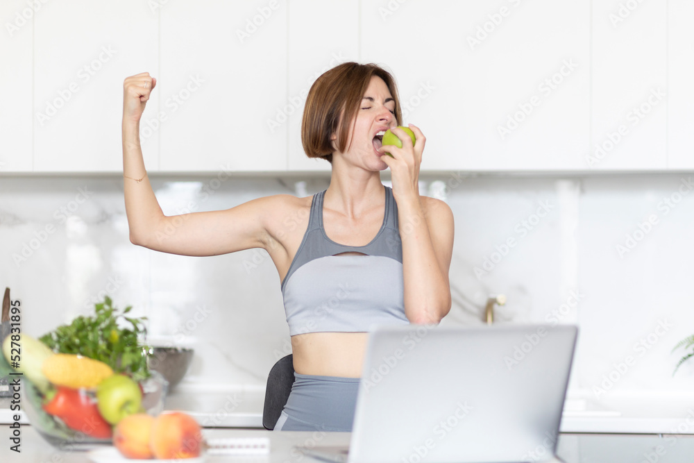 Young slender food blogger eating apple and demonstrating her biceps to her subscribers. Woman cooking healthy food using fruits and vegetables and biting apple at camera on laptop Young slender food blogger eating apple and demonstrating her biceps to her subscribers. Woman cooking healthy food using fruits and vegetables and biting apple at camera on laptop