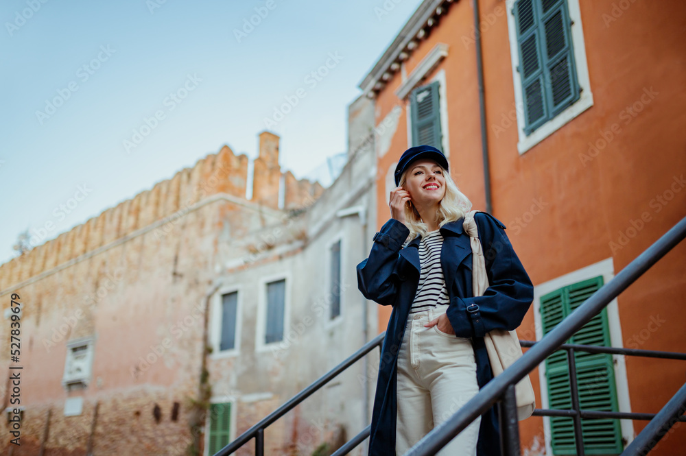 Happy smiling fashionable woman wearing trendy autumn outfit with baker boy cap, navy blue trench coat, striped shirt, white jeans, posing in street in Venice. Copy, empty space for text