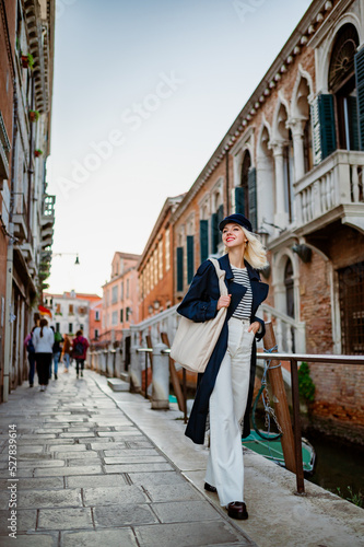 Happy smiling fashionable woman wearing trendy autumn navy blue trench coat, baker boy cap, striped shirt, white jeans, with beige leather hobo bag, walking in street in Venice © Victoria Fox