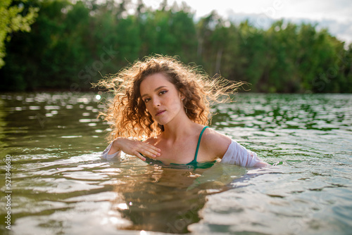 Beautiful curly girl in a white shirt swims in the lake. Girl in a wet white shirt. Green lake. Reserve. Girl resting on the beach 