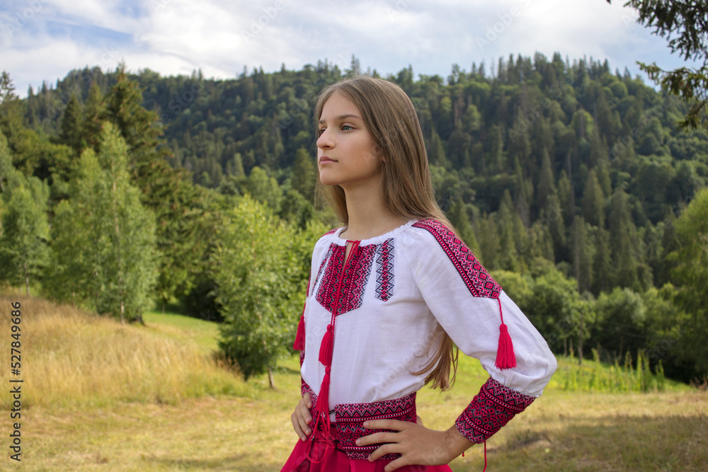 Obraz premium A young girl with loose hair in a national costume stands in profile in the mountains and looks straight ahead. Celebrates Independence Day of Ukraine. Cloudy summer weather in the Carpathians.