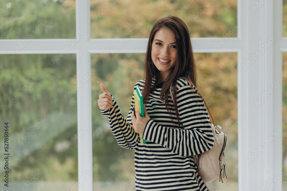 Happy cheerful beautiful high school, college or university student girl i isolated background, holding book and backpack, smiling and showing thumbs up Happy cheerful beautiful high school, college or university student girl i isolated background, holding book and backpack, smiling and showing thumbs up
