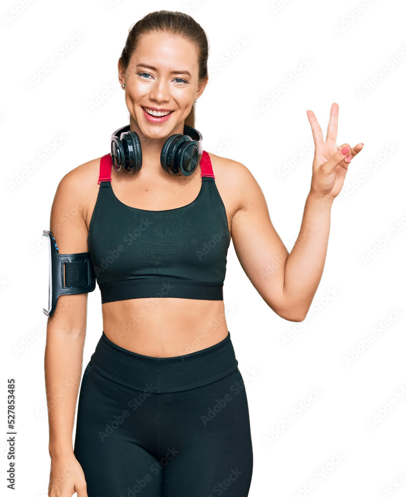 Beautiful blonde woman wearing gym clothes and using headphones smiling looking to the camera showing fingers doing victory sign. number two. Beautiful blonde woman wearing gym clothes and using headphones smiling looking to the camera showing fingers doing victory sign. number two.